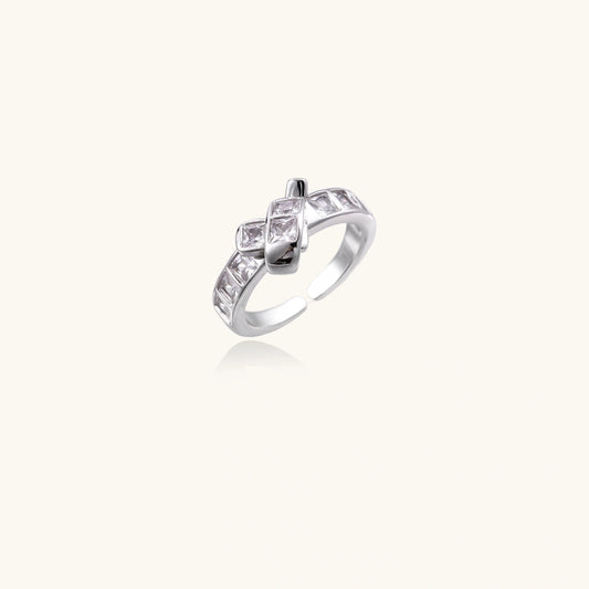 A silver-toned ring with a geometric, diamond-like pattern of clear stones, shown on a solid cream background with a soft reflection underneath.